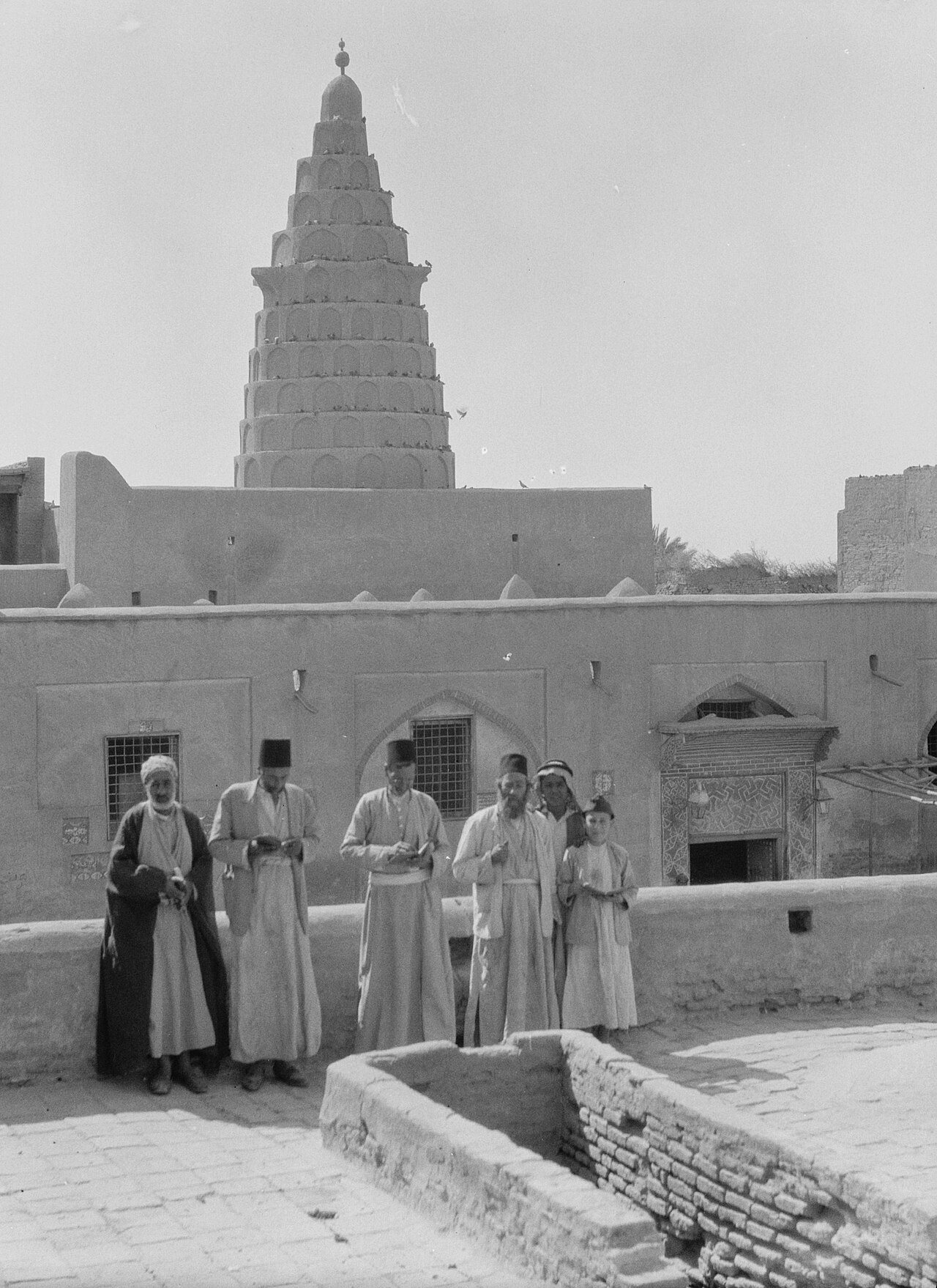 תצלום שחור לבן של גברים בגלביות ותרבושים, ומאחוריהם מגדל חרוטי מדורג