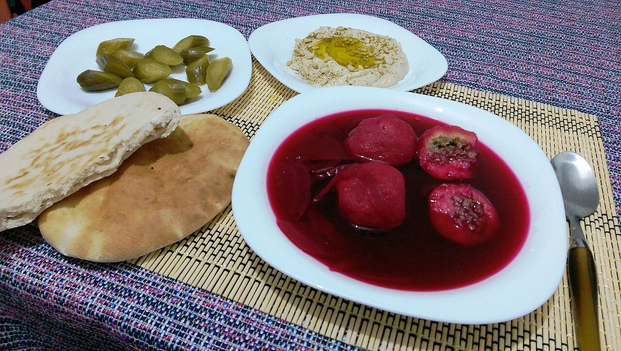 צלחת עמוקה עם מרק קובה סלק אדום, ולצדה פיתות, מלפפונים חמוצים וחומוס
