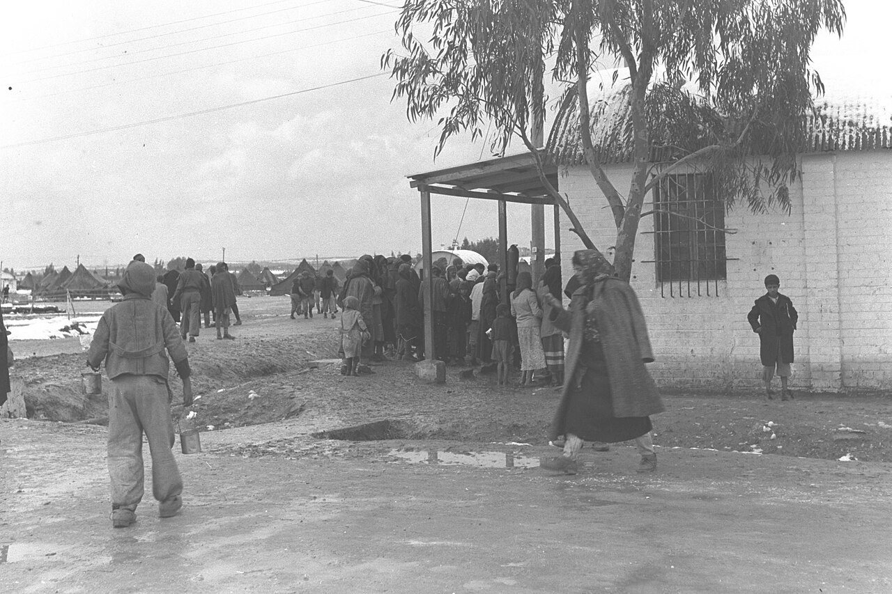 אנשים ממתינים ליד מבנה נמוך בדרך בוצית במחנה, ומאחור אוהלים