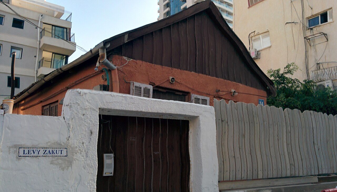 בית קטן וישן עם גג משופע, חומה לבנה ושער עץ, ומאחוריו בניינים גבוהים