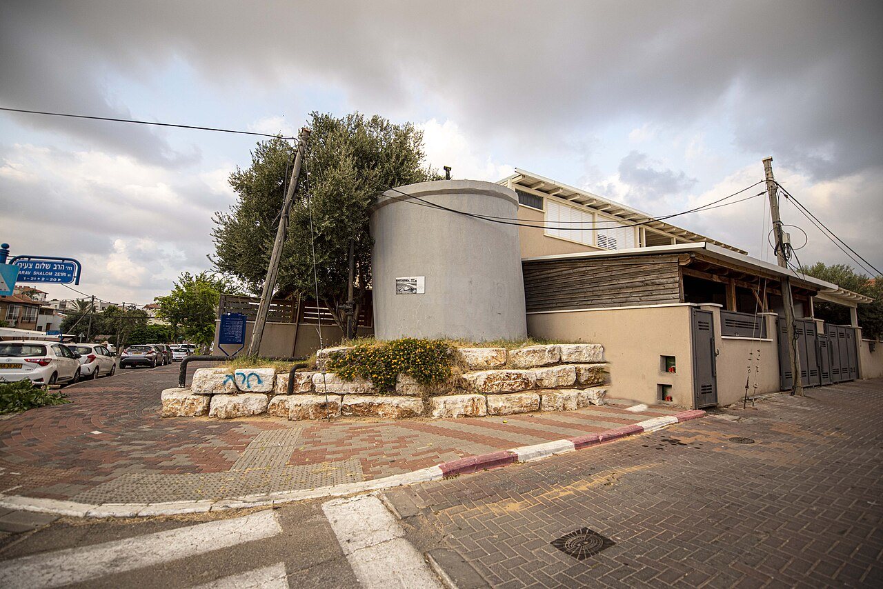 מבנה בטון גלילי על בסיס אבנים בפינת רחוב, לצד בית נמוך ועצים